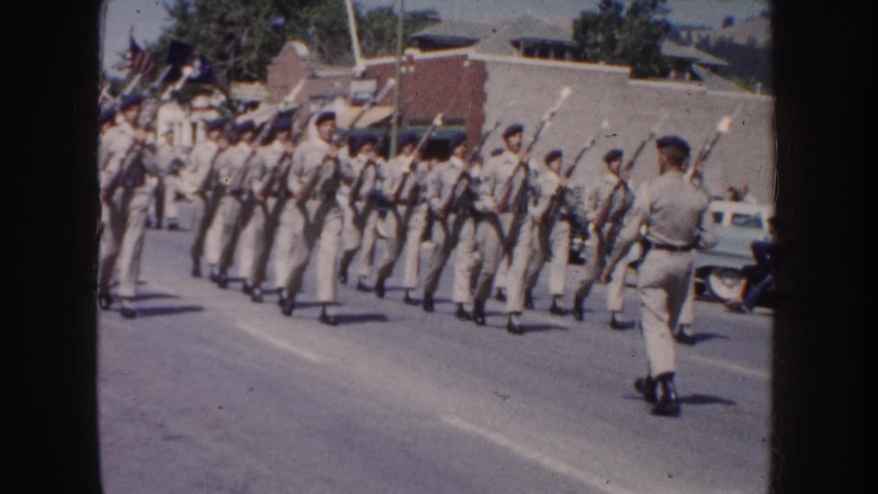 Uniformed marchers carry rifles while moving in formation past parked cars.