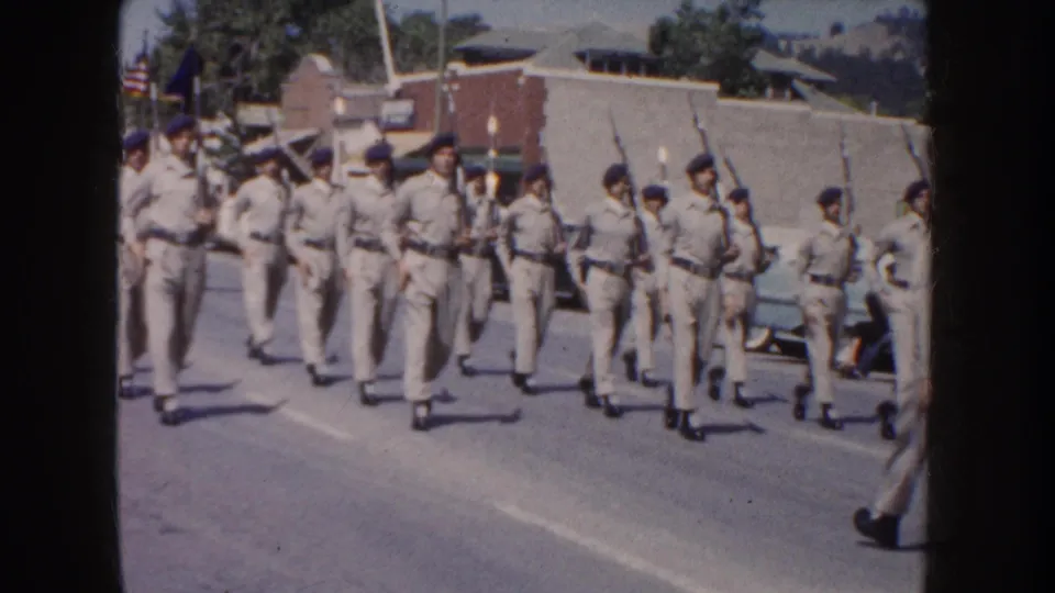 Roadside 8mm frame from a 1954 South Dakota parade showing cadets marching in step with rifles near parked cars.
