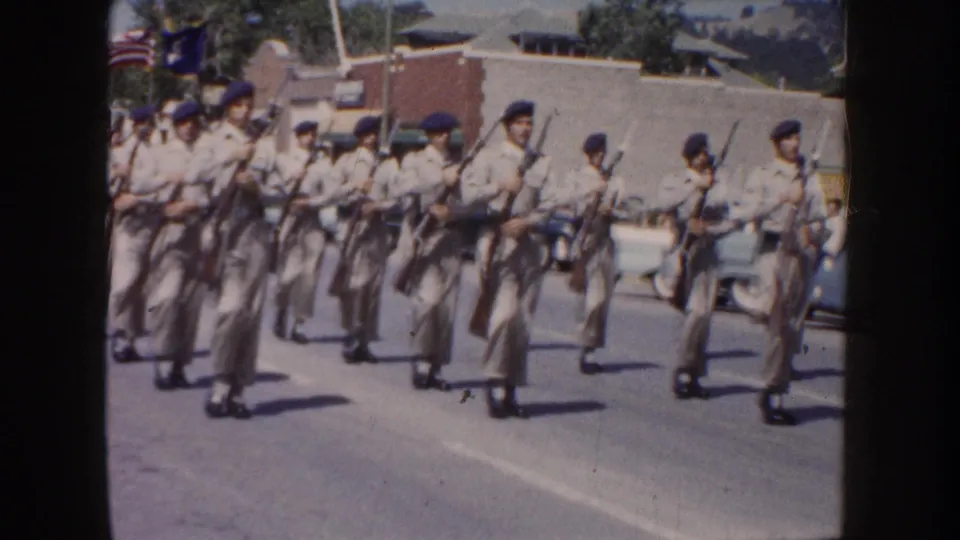 Close roadside view of 1954 South Dakota parade marchers in light uniforms carrying rifles through a town street.
