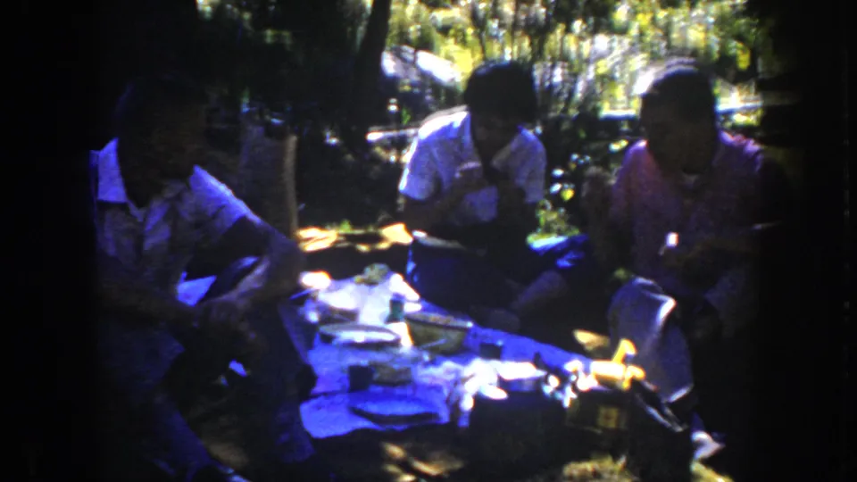 Three adults in casual 1950s clothing sit on a picnic blanket in South Dakota, eating and talking amid trees and grass.