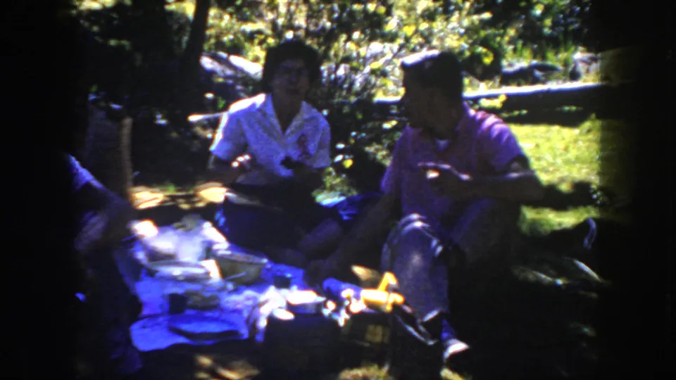 A clearer 1954 South Dakota frame shows friends seated beside picnic bowls and containers on a blanket in a wooded park.