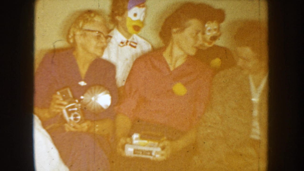 In the 1954 South Dakota home-movie frame, adults sit indoors near duck and puppet-like masks while one person holds a small device.