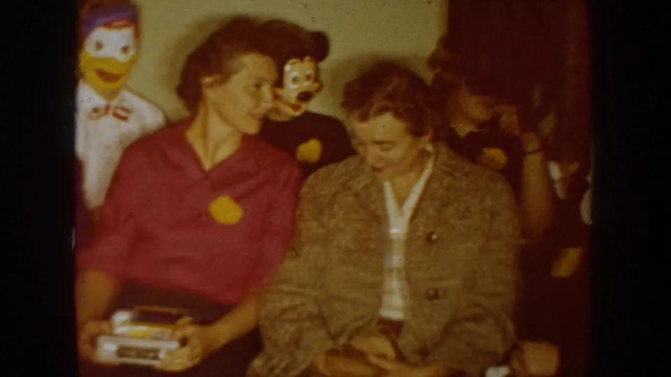 Two women sit closely in a 1954 South Dakota home-movie scene, with duck and puppet-like masks behind them and other adults nearby.