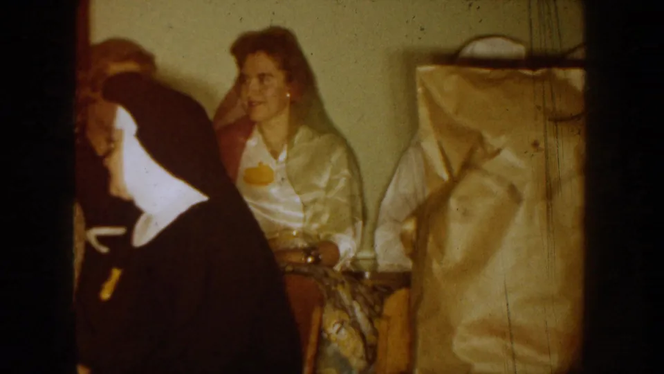 A woman seated indoors near a person in a nun-style habit appears in this 1954 South Dakota home-movie frame, with a hanging bag or garment at right.