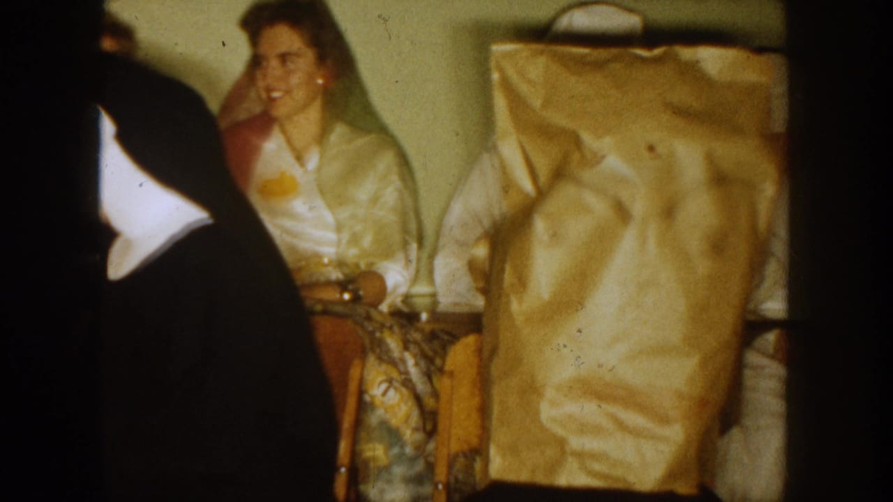 1954 South Dakota home-movie frame showing seated adults indoors, yellow paper bags over some heads, and a nun in black habit at left.
