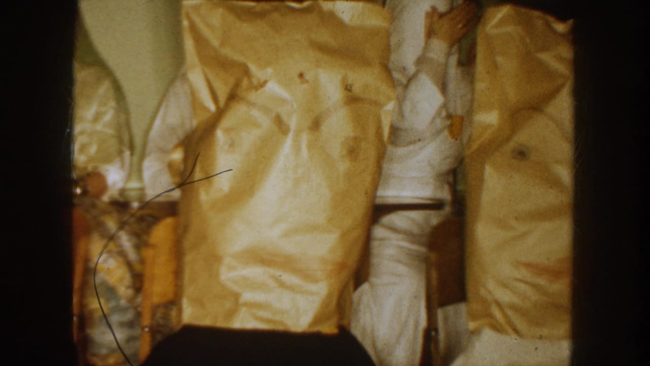 1954 South Dakota frame with seated adults in light clothing and yellow paper bags covering their heads inside a private-looking room.