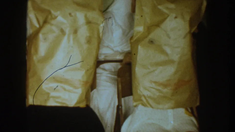 Close 1954 South Dakota home-movie view of seated figures with yellow paper bags over their heads and chair backs visible indoors.
