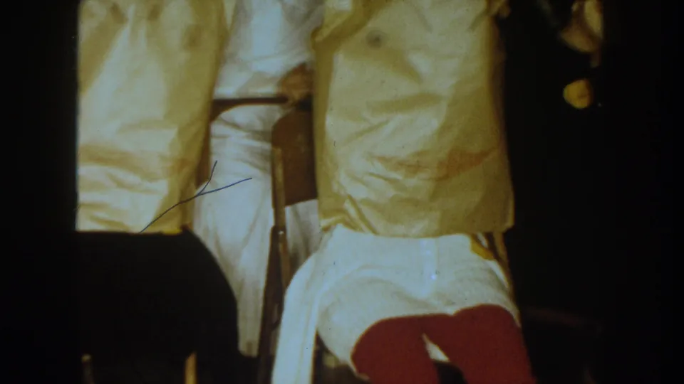 1954 South Dakota frame showing two seated paper-bag-covered figures, pale clothing, and a partial person at the right edge indoors.