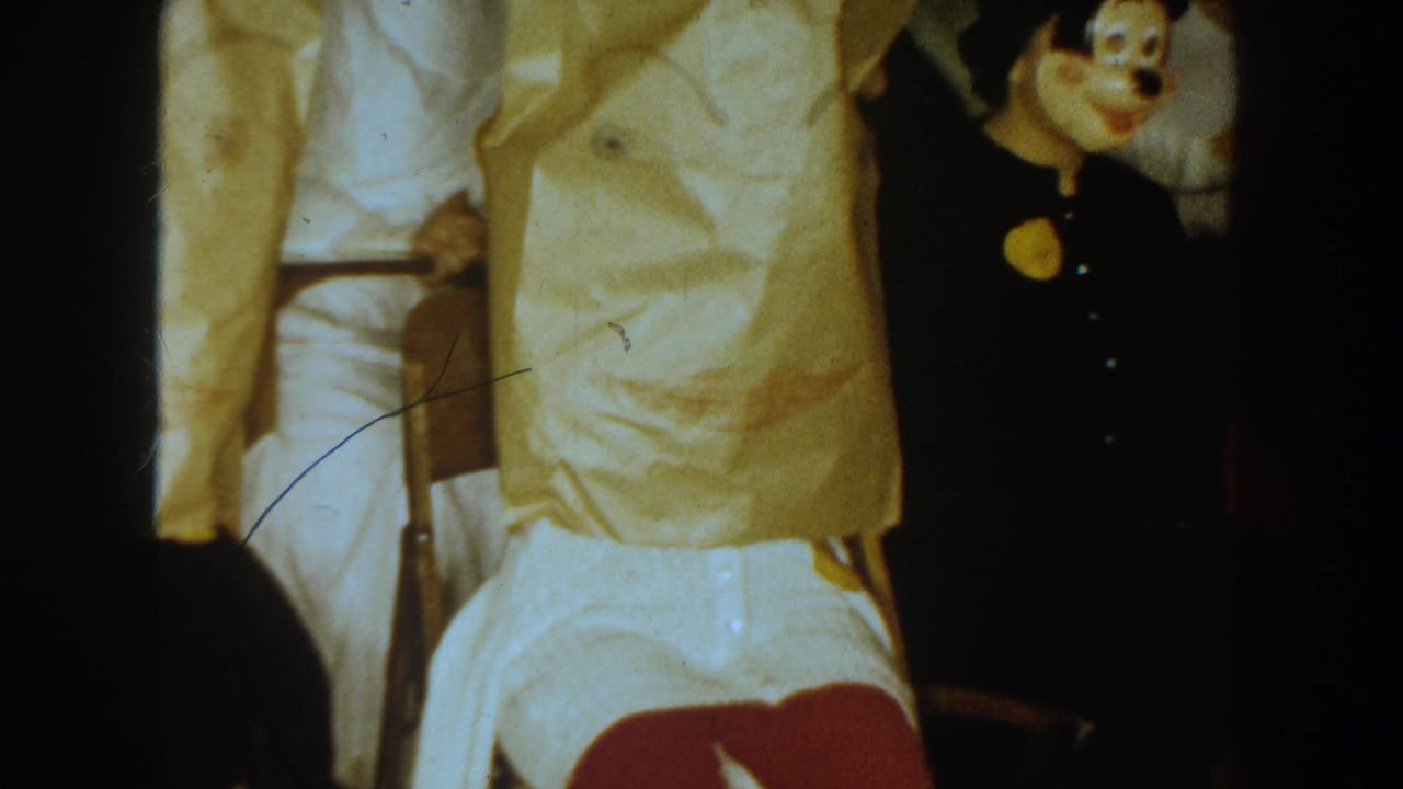 1954 South Dakota 8mm frame showing seated adults in costume, with a black cartoon mouse mask and pale clothing visible in low light.
