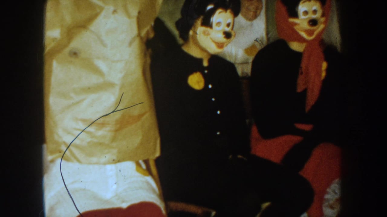 Two cartoon mouse masks appear among seated costumed people in a dark interior.