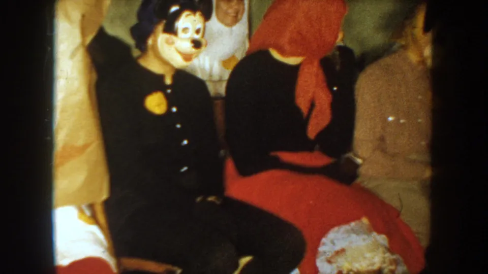 1954 South Dakota 8mm frame showing seated costumed adults, a black cartoon mouse mask, a red scarf, and period clothing.