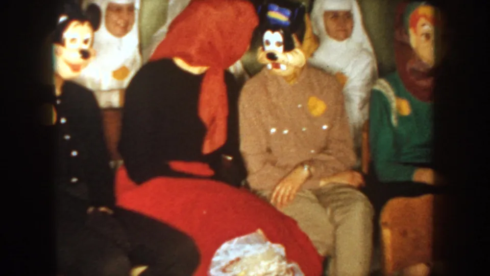 1954 South Dakota 8mm frame with adults seated in cartoon character masks, red scarf, brown jacket, and green garment visible.