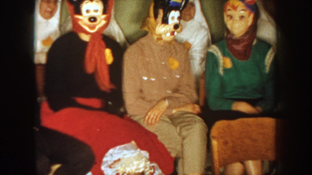1954 South Dakota home-movie frame showing seated adults in character-style Halloween masks, scarves, sweaters, and a private indoor party setting.