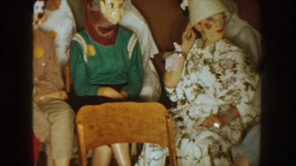 1954 South Dakota home-movie frame showing a woman in a floral dress and hat seated near adults in Halloween masks.