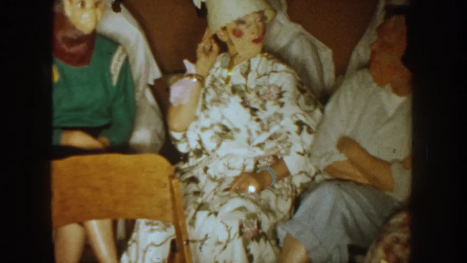 Final 1954 South Dakota frame with a floral-dressed woman, a seated unmasked adult, and nearby Halloween mask wearers in a private interior.