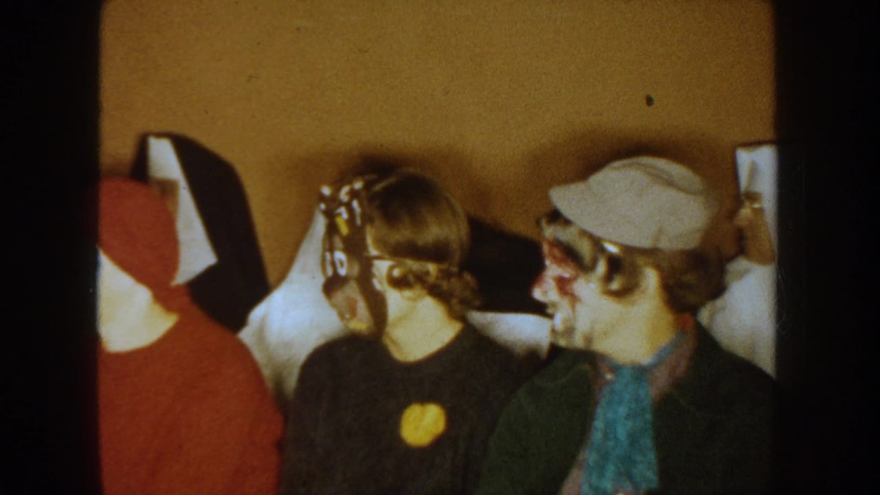 1954 South Dakota 8mm frame of adults seated close together indoors, including a red hood, mask, cap, and face paint.
