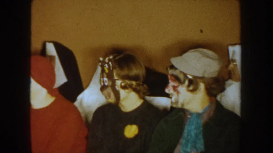1954 South Dakota 8mm frame of adults seated close together indoors, including a red hood, mask, cap, and face paint.