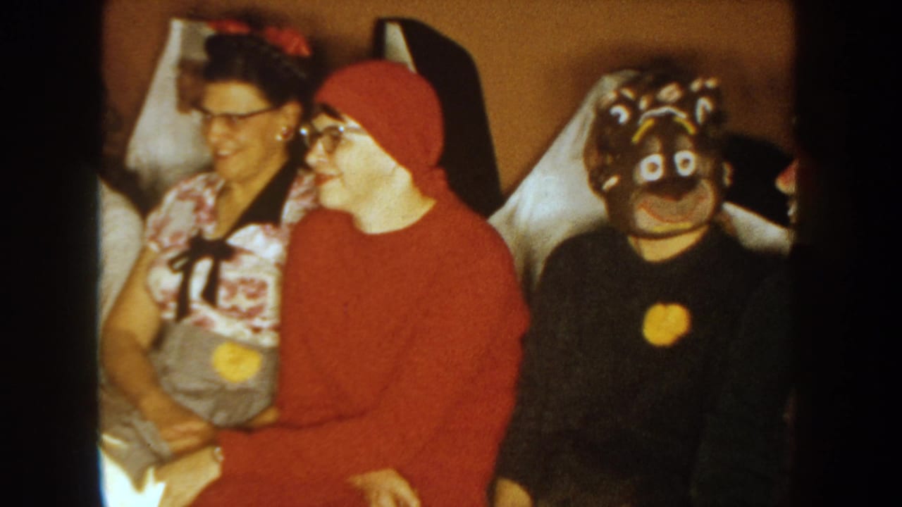 Three seated adults indoors, including one in a red hood and another in a dark mask.