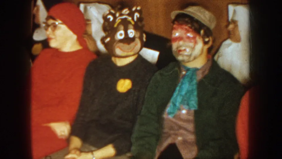 1954 South Dakota 8mm frame of adults seated at a masked gathering, with a red hood, dark mask, and painted face visible.