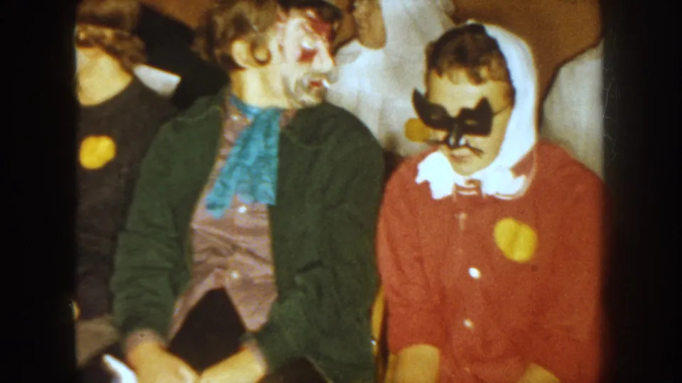 1954 South Dakota home-movie frame showing seated adults, including a painted face and a white hood with a dark mask.