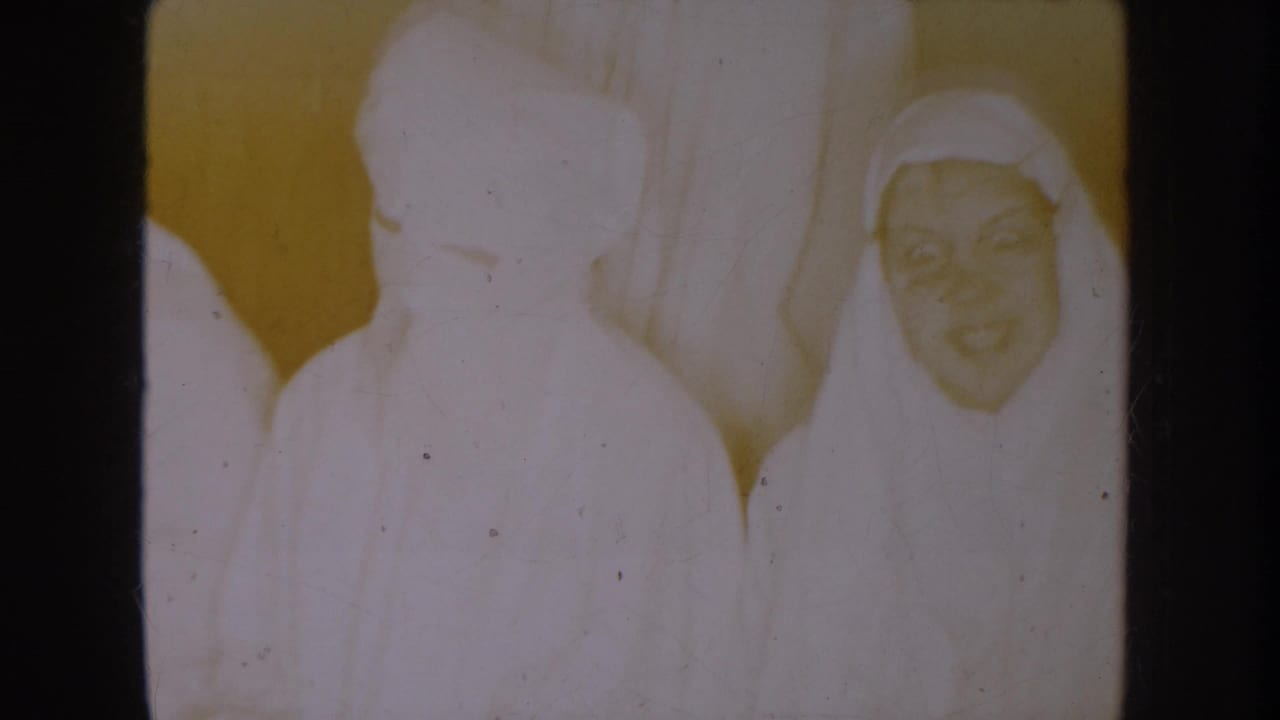 1954 South Dakota 8mm frame showing young figures in white nun-style robes and veils indoors, with one smiling face visible.