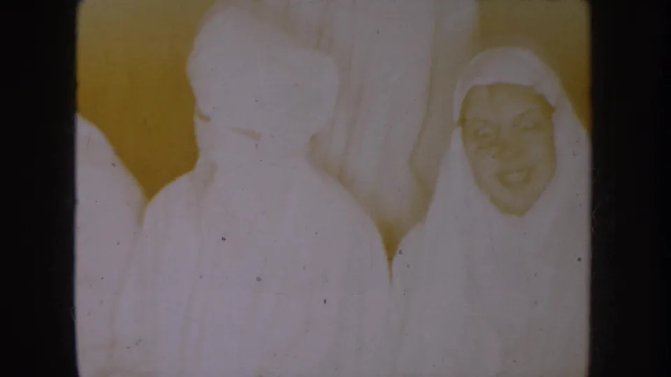 1954 South Dakota 8mm frame showing young figures in white nun-style robes and veils indoors, with one smiling face visible.