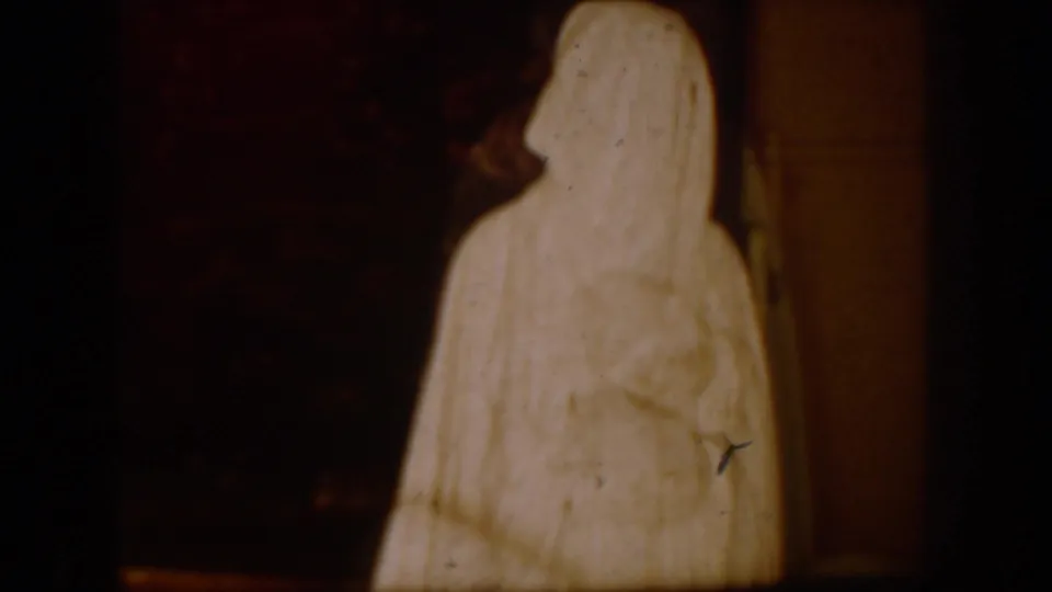 A tall figure in a white robe and veil appears indoors against a dark background in the 1954 South Dakota home-movie footage.