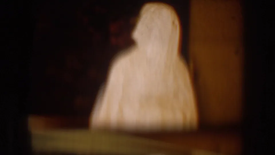 Soft-focused 1954 South Dakota 8mm frame of a white-robed, veiled figure standing in a dim private interior.