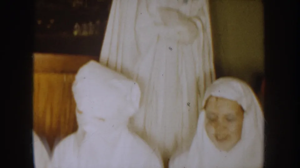 1954 South Dakota 8mm frame showing children in white veils near a taller robed religious figure in an indoor wood-paneled setting.