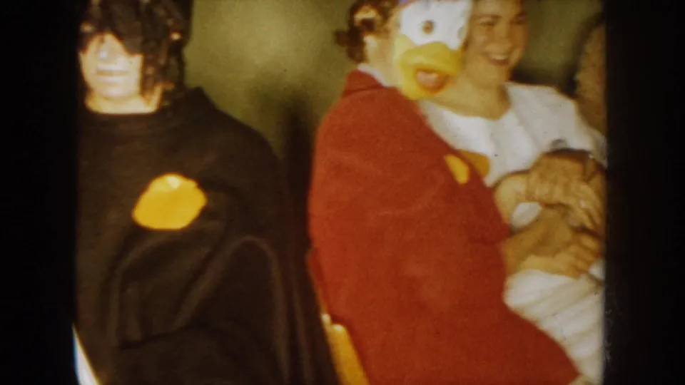 1954 South Dakota home-movie frame with party guests, including a yellow duck mask costume and a smiling person holding an infant.