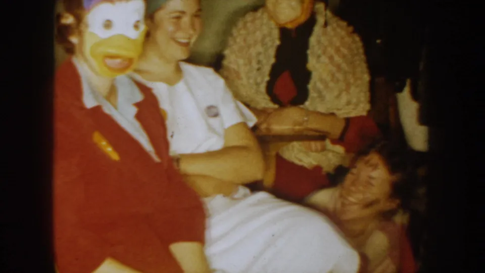 Warm 8mm frame of a South Dakota costume party with seated guests, a duck mask costume, and people smiling toward the camera.