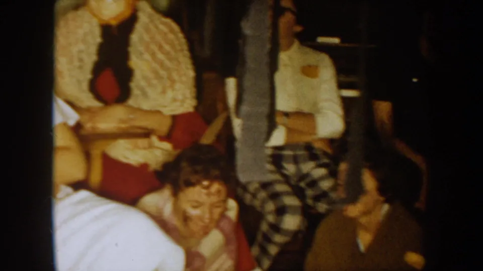 1954 South Dakota home-movie frame showing seated party guests in costume and patterned clothing inside a private interior.