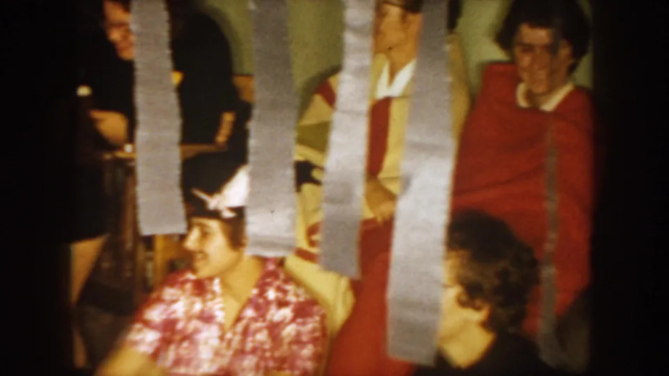 1954 South Dakota home-movie frame showing adults in an indoor party room, partly hidden by silver streamers.