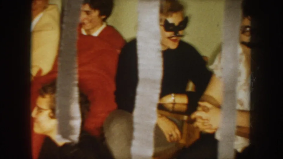 1954 South Dakota home-movie frame showing seated costume-party guests, including a masked adult, behind hanging streamers.