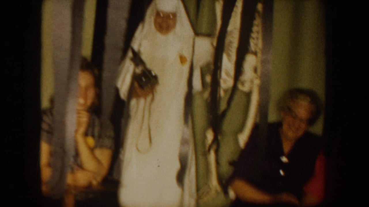 1954 South Dakota home-movie frame showing a woman in a white nurse-style uniform near seated adults in a dim private interior.