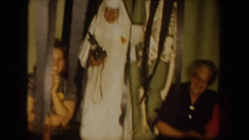 1954 South Dakota home-movie frame showing a woman in a white nurse-style uniform near seated adults in a dim private interior.