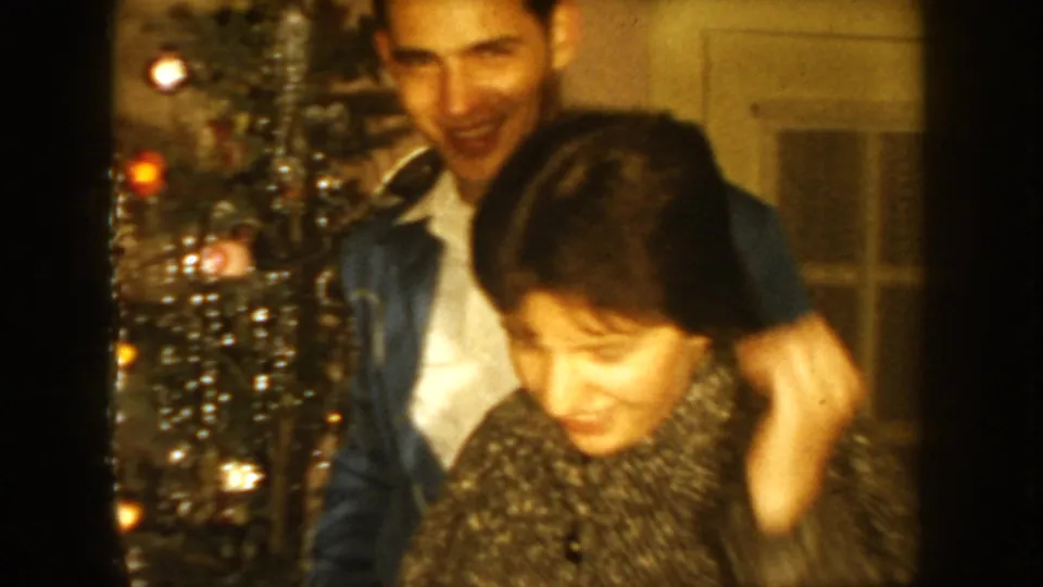 1954 South Dakota home-movie frame showing a smiling man in a denim jacket with a woman beside a lit Christmas tree indoors.