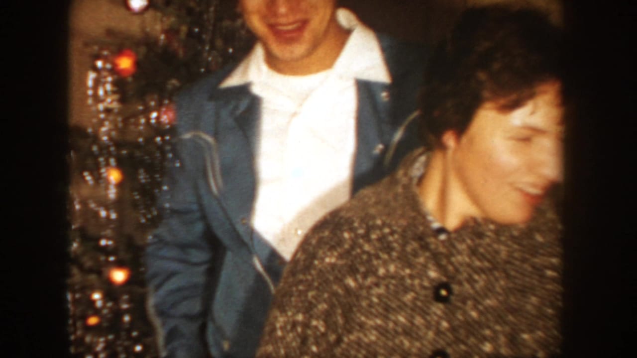 1954 South Dakota home-movie frame showing a smiling young man and woman beside a lit, decorated Christmas tree indoors.