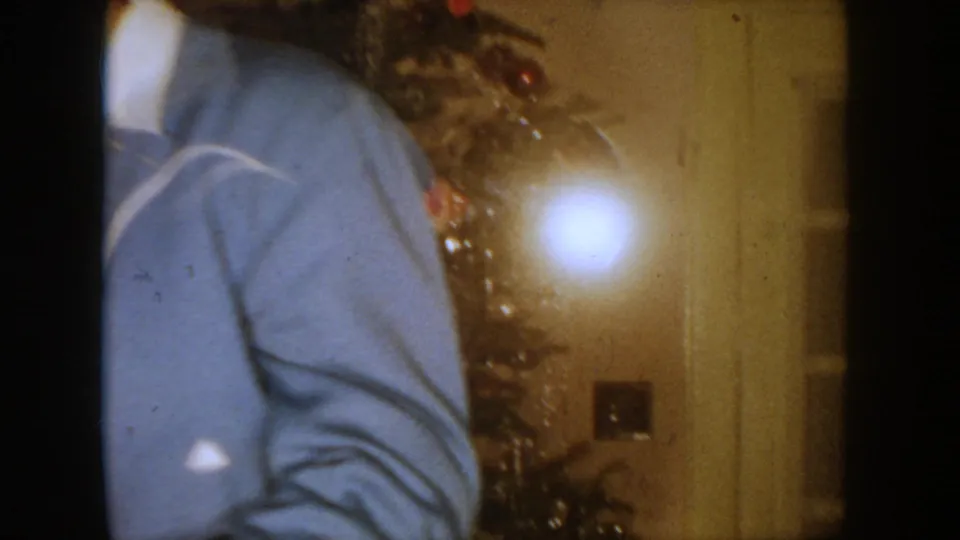 1954 South Dakota home-movie frame with a partial adult figure, decorated tree, and bright exposure flare inside a private room.