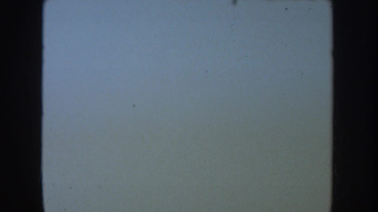1954 South Dakota 8mm reel frame showing a pale blue exposed field with black gate edge, grain, and scattered specks.
