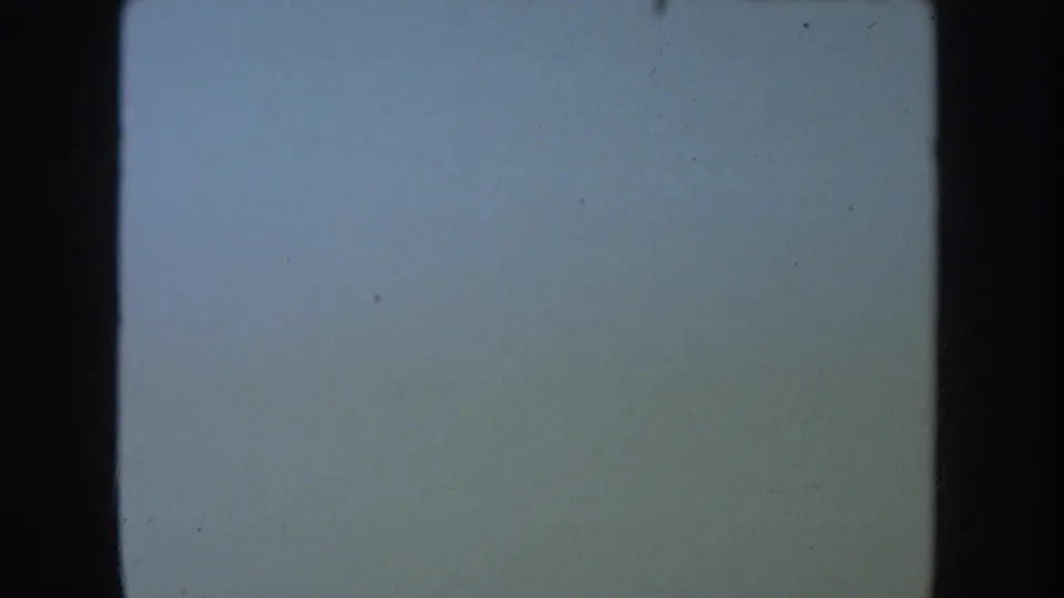 1954 South Dakota 8mm reel frame showing a pale blue exposed field with black gate edge, grain, and scattered specks.