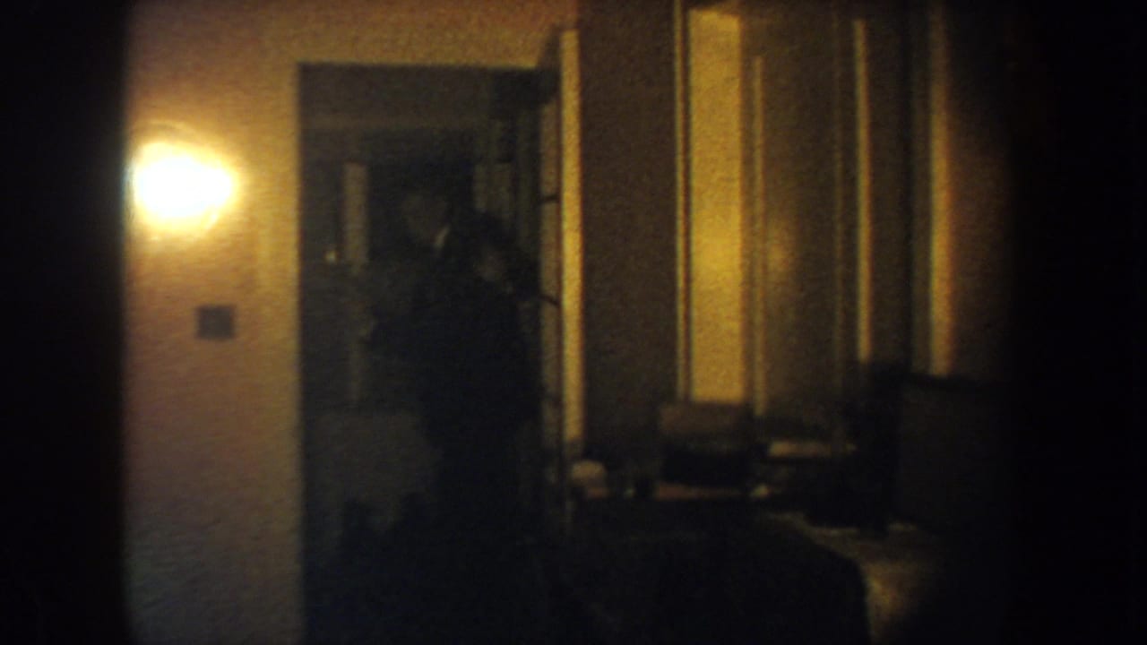 A man in a dark suit appears in a South Dakota home doorway, filmed in soft, grainy 8mm light.