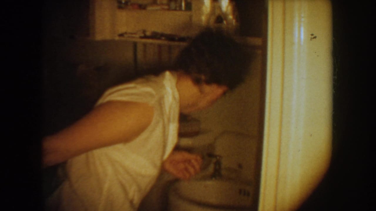 The woman bends or turns near the bathroom sink, framed through a doorway in grainy 8mm footage.