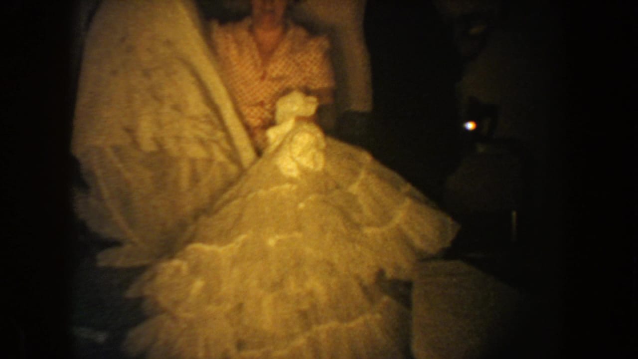 A woman in patterned indoor clothing holds a large pale frilled dress inside a dim private room in 1954 South Dakota footage.