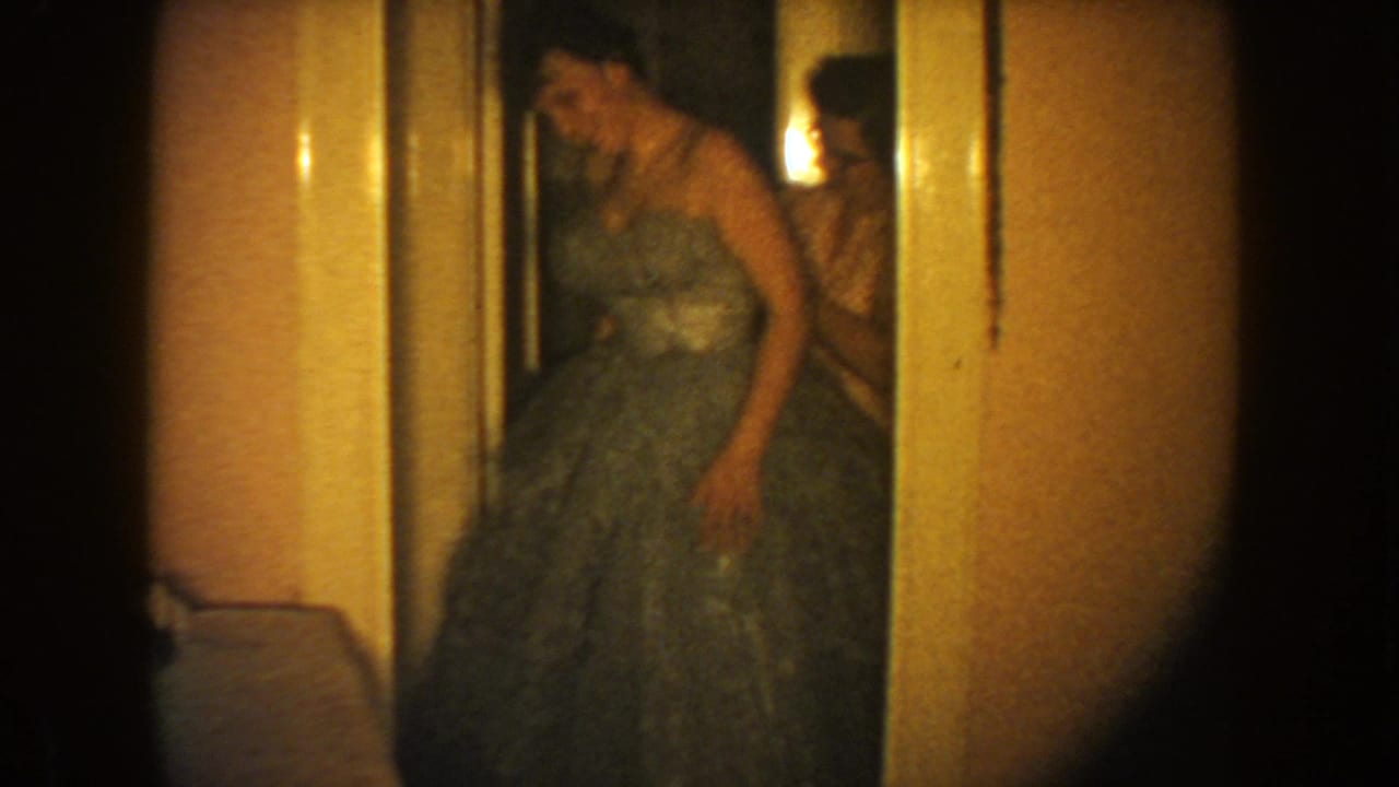 A woman in a strapless dark formal dress stands in a doorway, with another figure partly visible behind her in the dim interior.