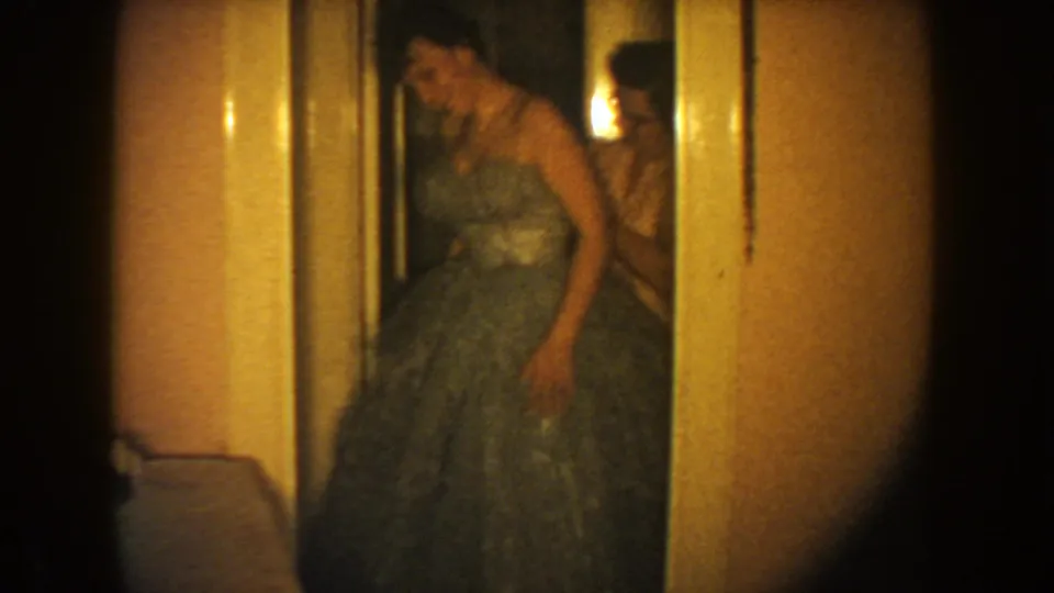 A woman in a strapless dark formal dress stands in a doorway, with another figure partly visible behind her in the dim interior.
