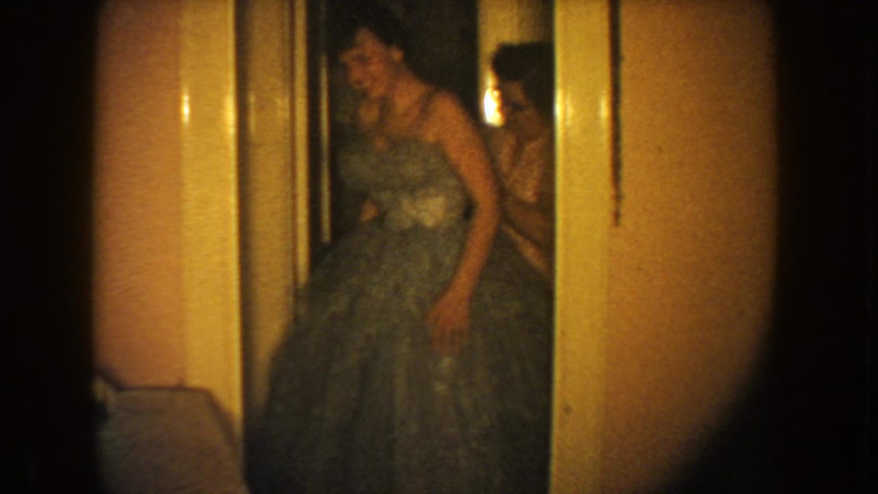 1954 South Dakota 8mm frame showing a woman in a full-skirted formal gown in a warmly lit hallway.