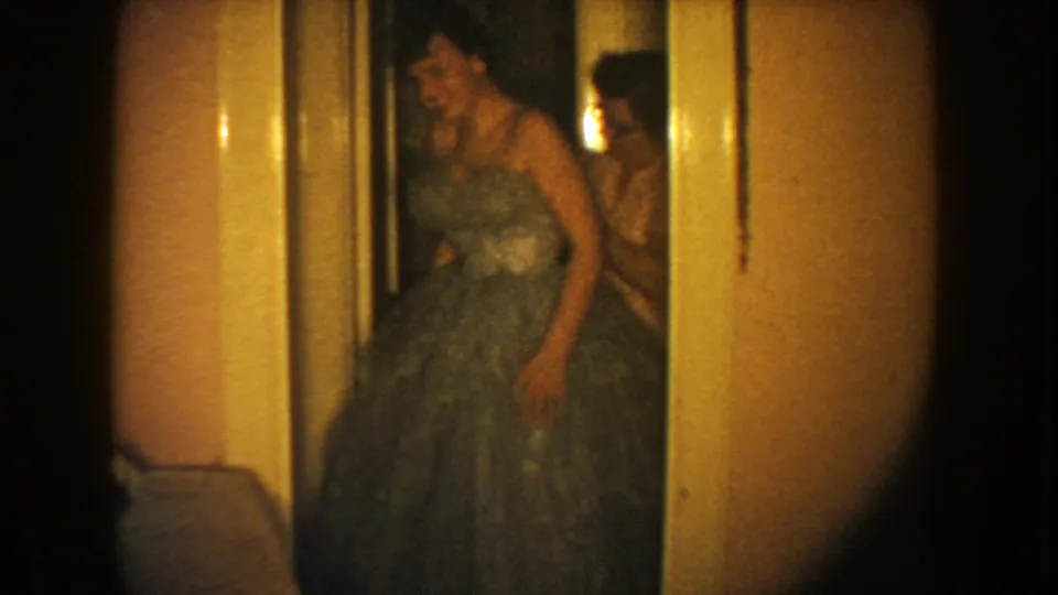 1954 South Dakota 8mm frame showing a woman in a full-skirted formal gown in a warmly lit hallway.