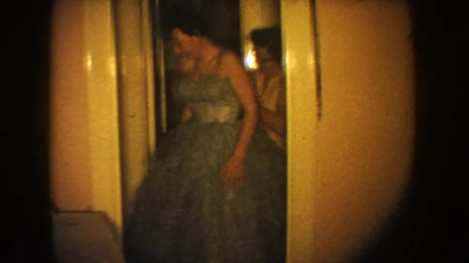 1954 South Dakota home-movie frame of a woman adjusting or holding a full formal gown in a dim interior.