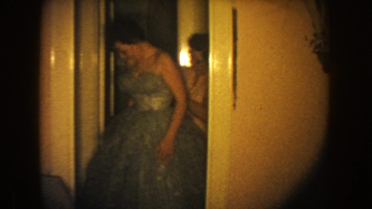 1954 South Dakota 8mm frame showing a woman in a full-skirted gown bending slightly in a private interior hallway.